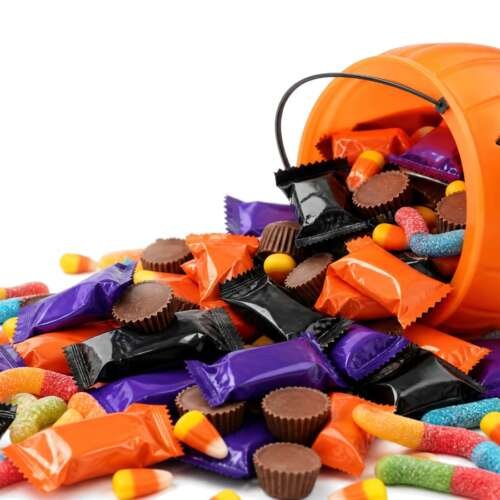 A tipped-over pumpkin trick-or-treat bucket spilling fun-size candy bars, peanut butter cups, candy corn and gummy worms on white
