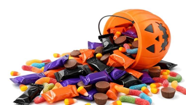 A tipped-over pumpkin trick-or-treat bucket spilling fun-size candy bars, peanut butter cups, candy corn and gummy worms on white