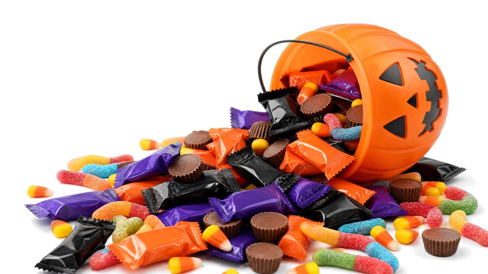 A tipped-over pumpkin trick-or-treat bucket spilling fun-size candy bars, peanut butter cups, candy corn and gummy worms on white