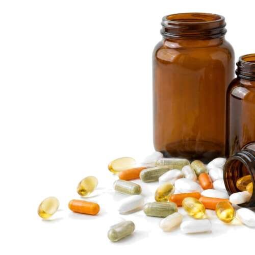 Assorted white and amber dietary supplement capsules spilling from a clear glass bottle on a bright white studio background
