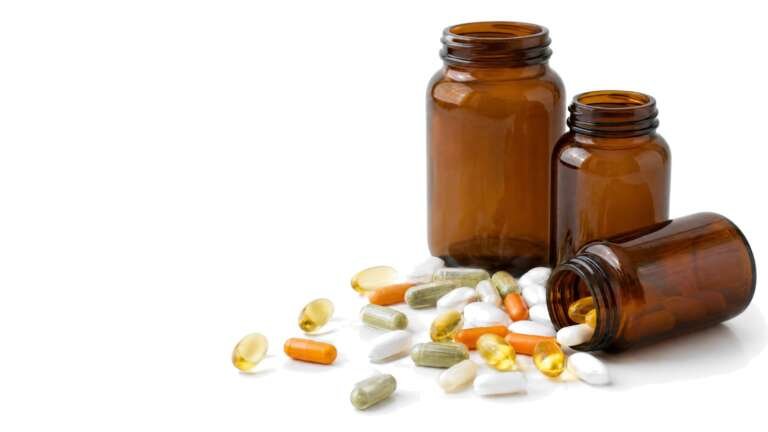Assorted white and amber dietary supplement capsules spilling from a clear glass bottle on a bright white studio background