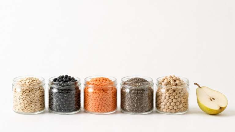Row of glass jars of oats, black beans, red lentils, chia seeds and chickpeas beside a halved pear