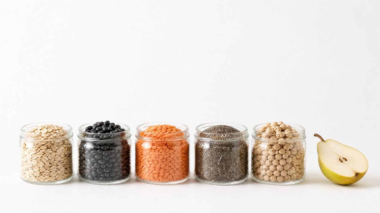 Row of glass jars of oats, black beans, red lentils, chia seeds and chickpeas beside a halved pear