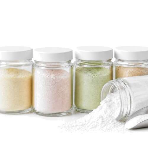 A row of small glass jars of colored ingredient powders on white, one tipped over with white powder spilling beside a steel scoop