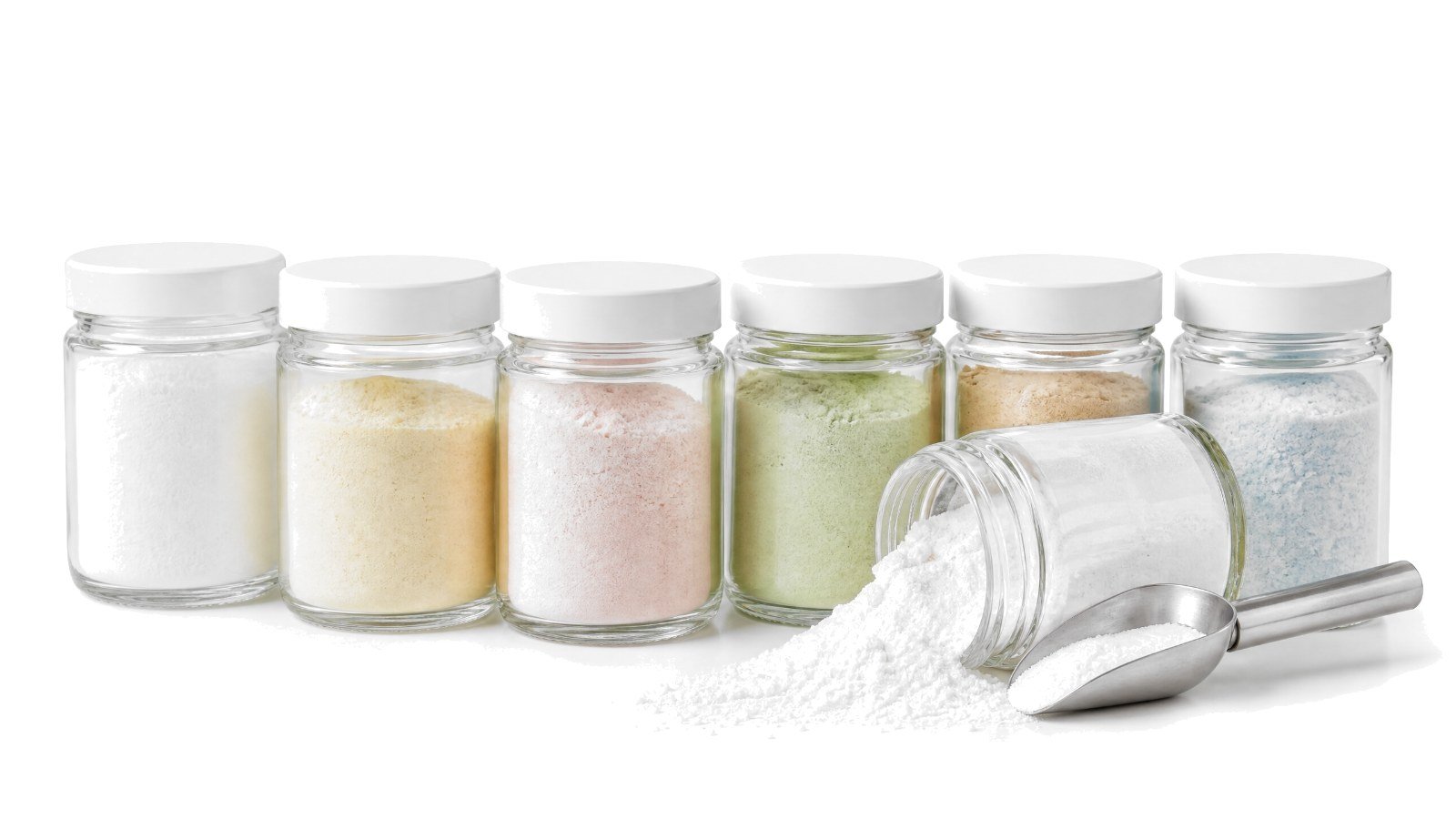 A row of small glass jars of colored ingredient powders on white, one tipped over with white powder spilling beside a steel scoop