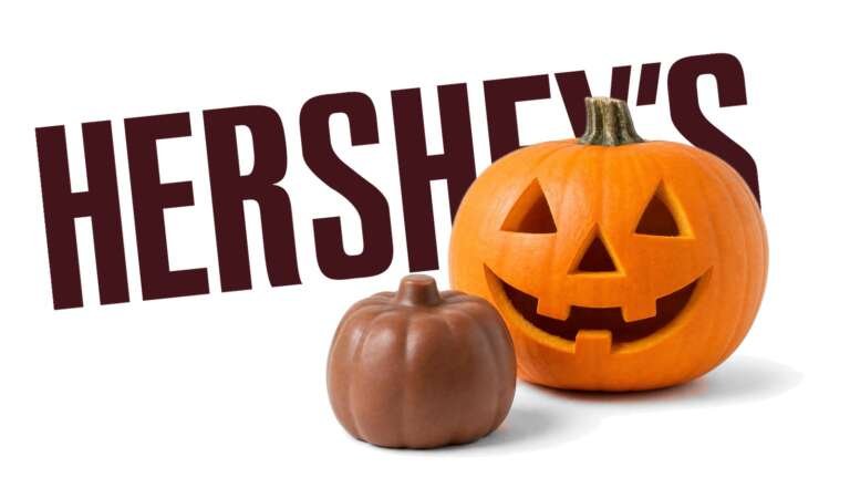 A carved jack-o'-lantern and a plain chocolate pumpkin candy in front of a large Hershey's logo