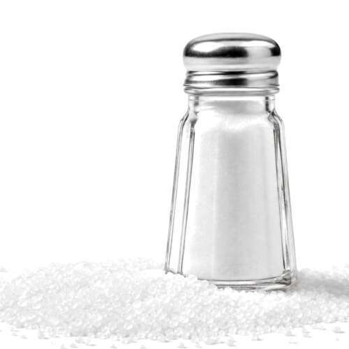A glass salt shaker standing in a spill of white salt crystals on a white background