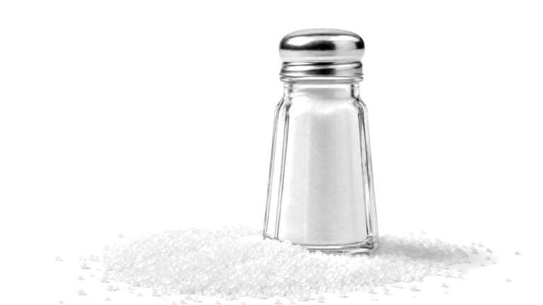A glass salt shaker standing in a spill of white salt crystals on a white background