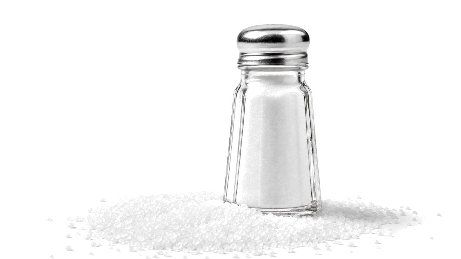 A glass salt shaker standing in a spill of white salt crystals on a white background