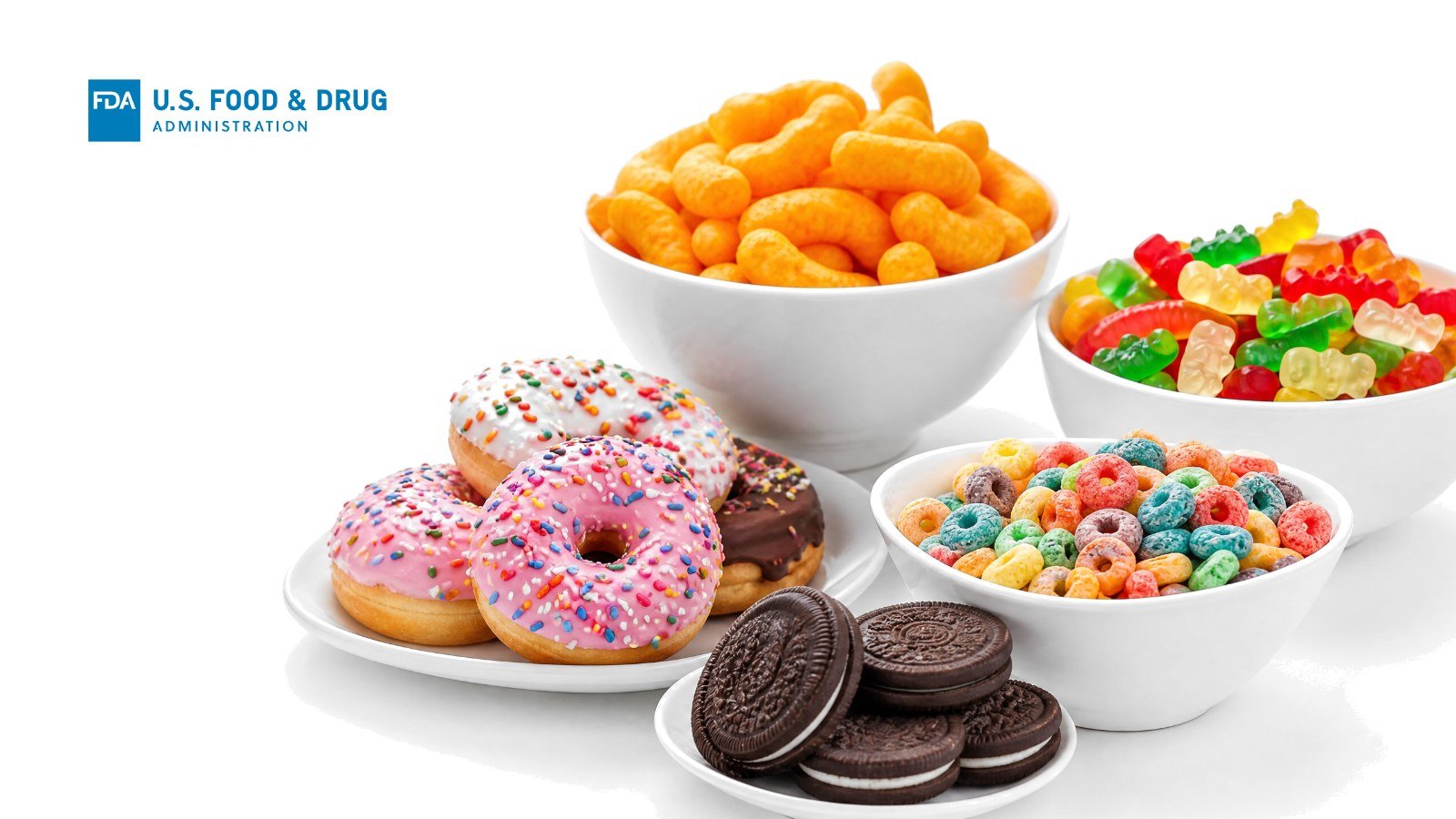Bowls of cheese puffs, gummy candy and frosted cereal loops with sprinkle donuts and sandwich cookies on white, with the FDA logo