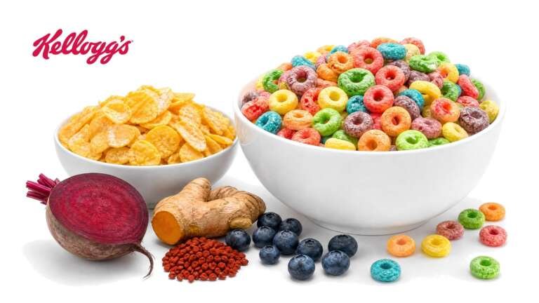 A bowl of brightly colored cereal loops and a bowl of frosted corn flakes with a halved beet, turmeric root, annatto seeds and blueberries on white, with the Kellogg's logo