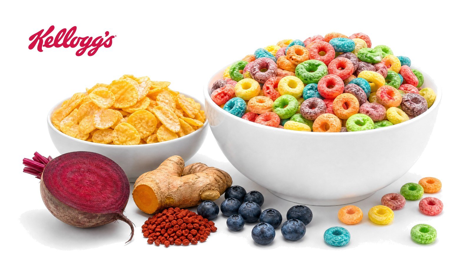 A bowl of brightly colored cereal loops and a bowl of frosted corn flakes with a halved beet, turmeric root, annatto seeds and blueberries on white, with the Kellogg's logo